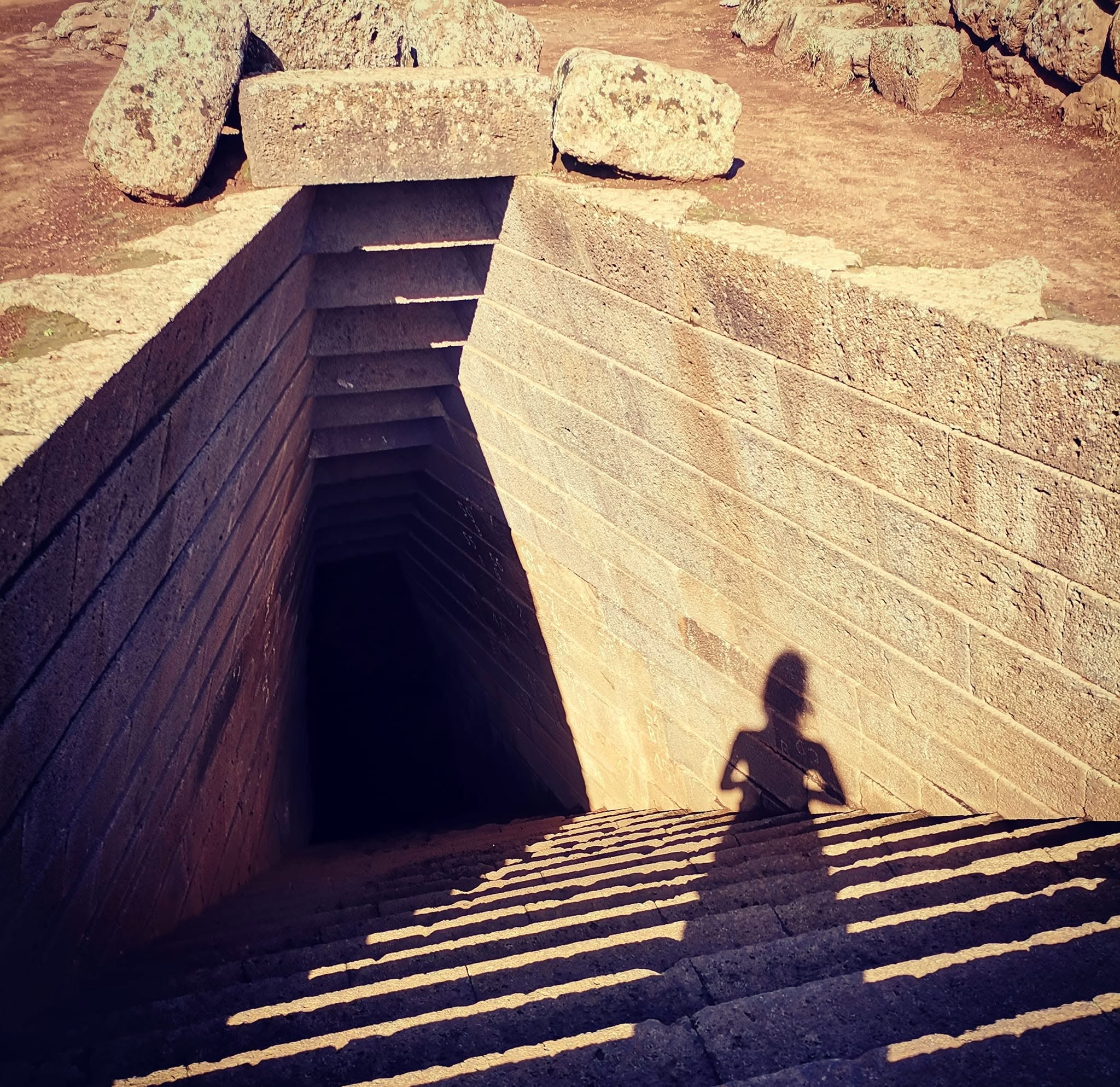 Ingresso in pietra di un'antica costruzione sotterranea con scalinata, l'ombra di una figura proiettata sulla parete